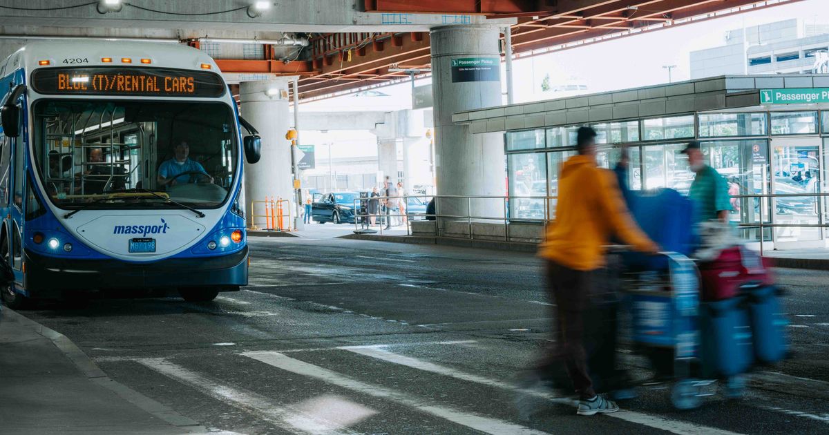 Busy airport scene showing travelers in motion and a rental car bus waiting under a covered terminal.