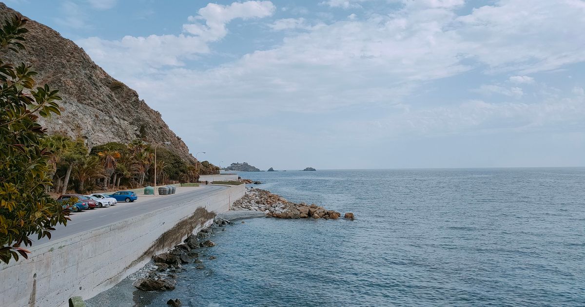 Serene view of a coastal road next to a calm ocean and rocky seashore under a partly cloudy sky.