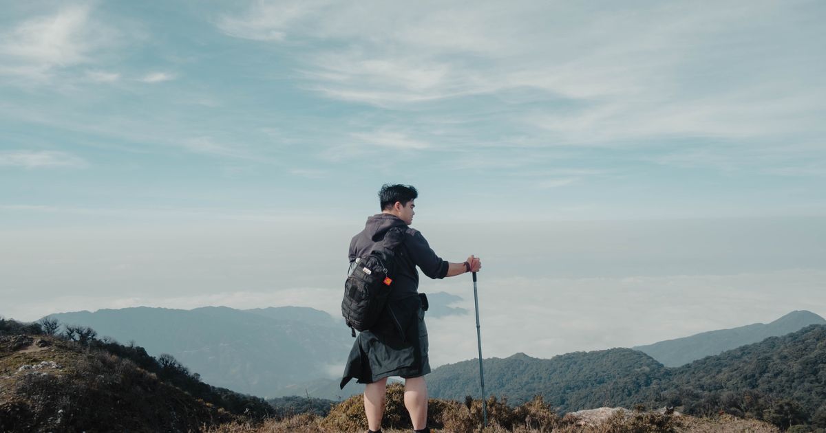 Solo hiker with backpack and walking stick looking at breathtaking mountain landscape under blue sky.
